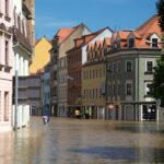 Street flooding in a European city with historic architecture under a sunny sky.