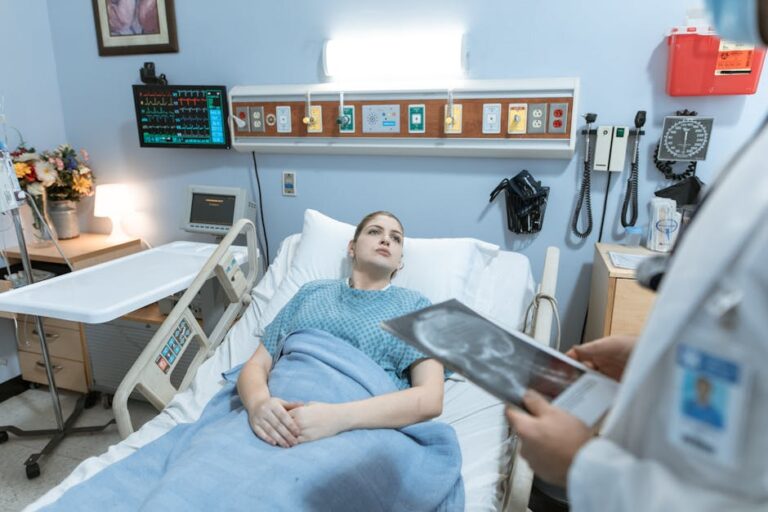 A female patient in a hospital gown receives medical attention from a healthcare professional holding an x-ray in a hospital ward.
