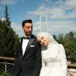 Bride and groom pose elegantly outdoors on a sunny day with a bridge backdrop.