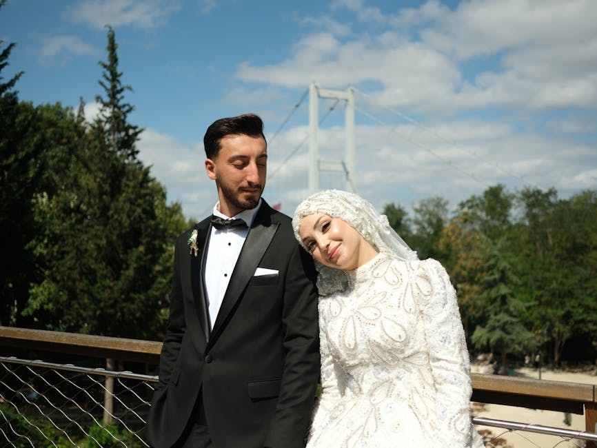 Bride and groom pose elegantly outdoors on a sunny day with a bridge backdrop.