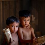Two young boys holding handmade bamboo instruments in a rustic wooden room.