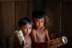 Two young boys holding handmade bamboo instruments in a rustic wooden room.