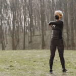 Full length young female in sportswear and mask making turns and kneading upper back while standing on green meadow in park