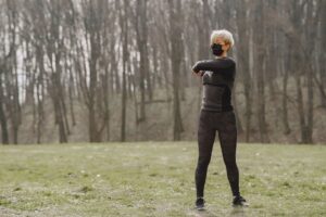 Full length young female in sportswear and mask making turns and kneading upper back while standing on green meadow in park