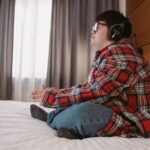 Young man with headphones and plaid shirt sitting comfortably in a bedroom, enjoying his leisure time.