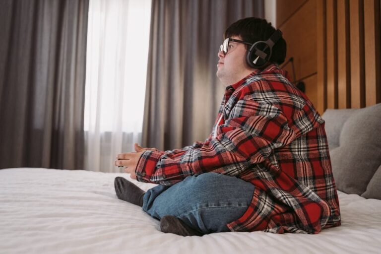 Young man with headphones and plaid shirt sitting comfortably in a bedroom, enjoying his leisure time.