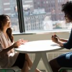 Two professional women having a discussion at a desk in a bright office setting.