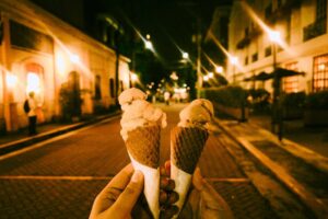 Two ice cream cones held up in a vibrant city street lit by warm streetlights at night.