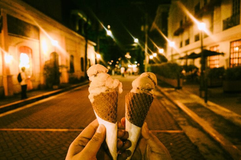 Two ice cream cones held up in a vibrant city street lit by warm streetlights at night.