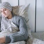 Woman resting in bed wearing a scarf, using her phone during cancer treatment.
