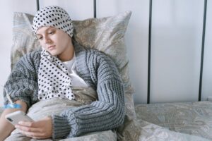 Woman resting in bed wearing a scarf, using her phone during cancer treatment.
