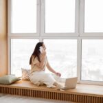 A woman talking on a smartphone while sitting by a large window with a laptop. A cozy indoor setting.