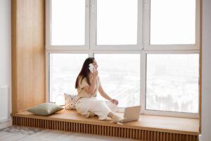 A woman talking on a smartphone while sitting by a large window with a laptop. A cozy indoor setting.