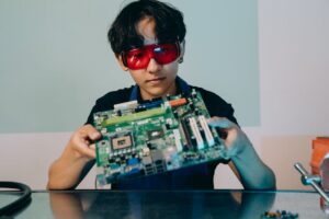 A focused young engineer holding a motherboard, wearing eye protection indoors.