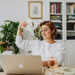 Smiling woman holding cash while working on a laptop in a cozy home office setting.
