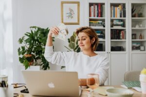Smiling woman holding cash while working on a laptop in a cozy home office setting.