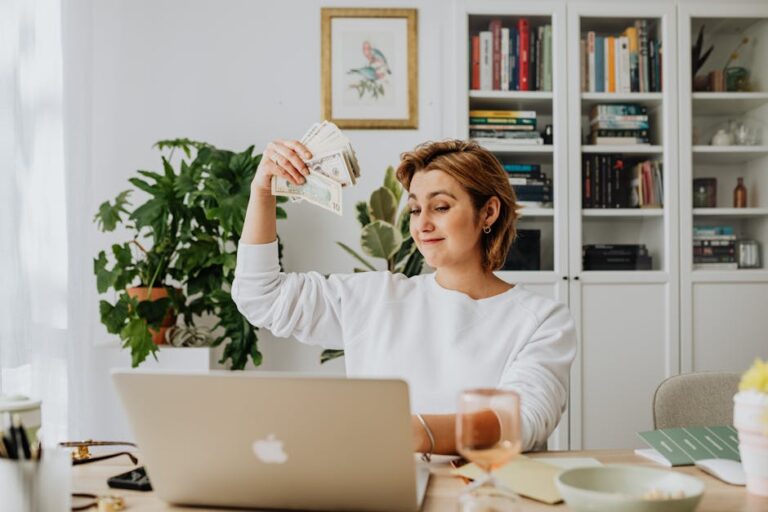 Smiling woman holding cash while working on a laptop in a cozy home office setting.