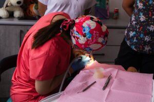 A pediatric dentist wearing a cartoon cap treats a child in a dental clinic.